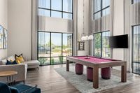 Elegant game room with high ceilings, featuring a stylish purple pool table and cozy seating, enhanced by floor-to-ceiling windows providing abundant natural light.