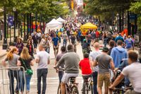 Vibrant city street filled with people walking and cycling, showcasing a lively and engaging community event under green tree canopies and colorful tents.