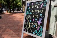 Charming sidewalk chalkboard with colorful floral designs and uplifting 'Here Comes the Sun' message.