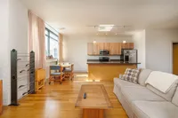 Bright and modern open-plan living area featuring a minimalist kitchen with wooden cabinetry, cozy lounge with a comfortable beige sofa, and a sunlit dining space near large windows.