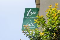 Inviting green sign for Trio Townhomes North against clear sky, surrounded by vibrant green foliage.