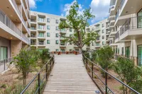 Modern apartment complex featuring lush greenery and a serene wooden walkway leading to outdoor seating beneath a towering tree.