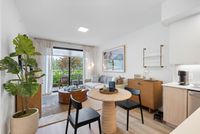Bright and modern open-concept living and dining area with stylish decor, featuring a cozy sofa, elegant artwork, and lush greenery with a view through large glass doors.