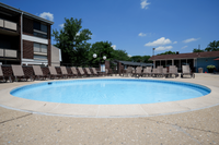 Charming apartment complex featuring a refreshing circular swimming pool surrounded by comfortable lounge chairs, set against a backdrop of lush trees and a clear blue sky.