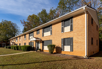 Charming brick apartment building with classic shutters and manicured landscaping, set against a backdrop of vibrant trees and a clear blue sky.