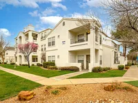Charming apartment complex surrounded by lush greenery and bright blossoming trees, featuring crisp white architecture under a clear blue sky.