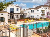Charming apartment complex with a pristine pool, sun loungers, and vibrant outdoor seating, surrounded by elegant white buildings under a clear blue sky.
