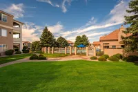 Charming apartment complex with lush green lawns, tall trees, and a sparkling swimming pool under a clear blue sky.