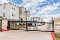 Gated entry to a modern apartment complex with elegant stone facade and ample parking, set against a clear blue sky.