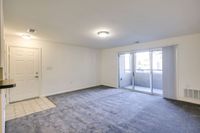 An unoccupied room with plush carpet and a sliding glass door, situated in Allyson Gardens Apartments II.