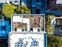 Aerial view of a modern rooftop garden with lush greenery and elegant seating area, surrounded by urban architecture and vibrant trees.