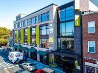 Modern urban building with large windows and vibrant green accents, featuring a prominent 'Now Leasing' sign for luxury apartments. Bustling street view with parked FedEx truck, adding to the lively neighborhood atmosphere.