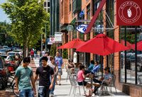 Vibrant urban street scene with people enjoying a sunny day at outdoor cafes under bright red umbrellas. Charming brick storefronts and lush green trees line the bustling sidewalk.