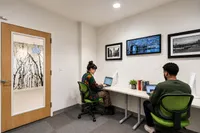 Modern co-working space featuring two people working at laptops on a sleek white desk with vibrant green chairs, contemporary art on the walls, and ample lighting.