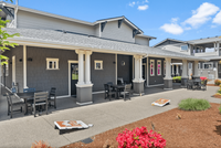 Relax and unwind in this inviting outdoor patio, complete with comfortable seating and fun lawn games. Outdoor patio area with tables, chairs, and cornhole boards in front of a modern gray building with white trim and flowering bushes.