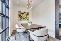 A modern conference area features a wooden table surrounded by two white upholstered chairs, with a vibrant abstract painting on the wall and a small potted plant as a centerpiece. Natural light streams in through large windows, creating a bright and inviting atmosphere.