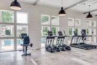 Bright fitness room featuring exercise equipment including treadmills and an elliptical, with large windows providing natural light and a view of the outdoor pool area.