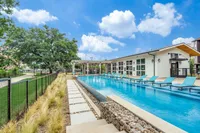 A clear blue swimming pool with lounge chairs is surrounded by a well-maintained landscape, featuring greenery and a modern building in the background. The sky is bright with fluffy clouds.