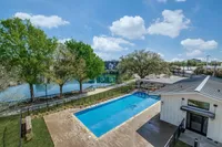 A clear view of a swimming pool surrounded by greenery, with a building nearby and a serene lake in the background under a bright blue sky.