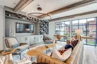 A modern lounge area featuring a stylish sofa, wooden tables, and a large TV against a decorative wall. Large windows provide natural light and a view of the outdoors.