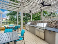 An outdoor grilling area features stainless steel barbecue grills, a stone wall, and modern seating under a pergola. Lush greenery and a blue sky create a relaxing atmosphere.