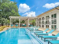A sparkling outdoor pool surrounded by lounge chairs and shaded areas, with a modern building in the background and a clear blue sky above.