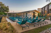A modern outdoor pool area features lounge chairs arranged around a sparkling blue pool, with a clubhouse in the background and landscaped greenery nearby. The setting is bright and inviting, perfect for relaxation.