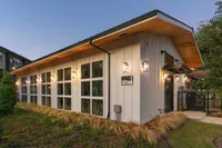 A modern building with large windows and a wooden overhang, surrounded by grassy landscaping. The exterior features a mix of white and natural wood elements, creating a welcoming atmosphere.
