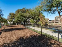 Fenced dog park with mulch and trees alongside a fenced path near a calm body of water. Buildings are visible in the background under a clear blue sky.