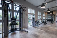 A modern gym interior featuring various exercise equipment, including a cable machine and weight benches, with large windows allowing natural light. The space has a clean and open design.