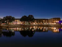 A serene lakeside view at dusk, showcasing buildings and trees reflected in the calm water. The scene is illuminated by soft lights from the nearby structures.