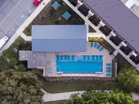 Aerial view of a swimming pool area surrounded by lounge chairs and a nearby building. The pool is rectangular, with a shaded area and green landscaping around it.