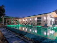 A modern building with large windows is illuminated at dusk, featuring a swimming pool with green lighting and lounge chairs around it. The serene setting includes a patio area with a pergola.
