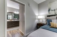 Cozy bedroom with plush bedding and elegant lamp, leading into a stylish bathroom with modern cabinetry and a spacious mirror.