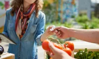 person holding tomatoes and giving credit card to woman at outdoor shop on sunny day