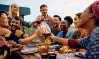 Vibrant group of friends toasting at an outdoor table with food and drinks, enjoying a sunny day.
