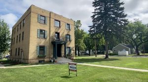Charming three-story brick apartment building with classic architecture, lush green lawn, and cozy outdoor seating. Ideal location for vibrant living surrounded by tree-lined streets.