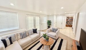 Bright and inviting living room with cozy white sofas, stylish striped cushions, and a chic wooden coffee table adorned with plants, leading to a tiled entryway with built-in shelves.