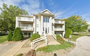 Charming three-story white apartment building with spacious balconies, surrounded by lush green trees and a clear blue sky.