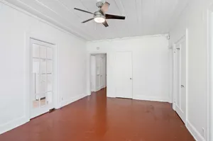 Bright, freshly painted room with polished wooden floors and a sleek modern ceiling fan, offering a glimpse into an adjacent kitchen through a stylish glass door.