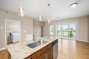 Bright apartment interior featuring a modern granite countertop island, stylish pendant lighting, open-concept living area, and large sliding glass doors leading to a balcony with scenic views.