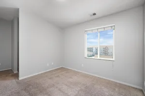 Bright and airy bedroom with soft carpet flooring and a large window offering a stunning view of neighboring buildings under a clear blue sky.