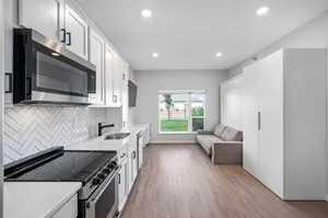 Bright modern kitchen with sleek white cabinets, herringbone backsplash, and stainless steel appliances, leading to a cozy living area with large window views.