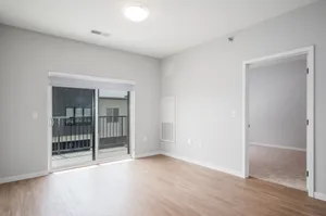 Bright empty room featuring beautiful wood flooring, large sliding glass doors leading to a balcony, and a cozy adjacent carpeted space.