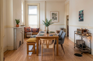 Charming dining area with stylish wooden table set for four, surrounded by cozy benches and chairs. Accents include a lush plant centerpiece, decorative pillows on a wooden bench, and warm lighting on a vintage-style bar cart.