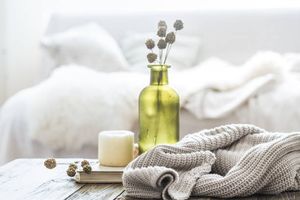 Cozy bedroom scene with a green vase, soft knits, and ambient candle on a rustic wooden table, inviting relaxation.