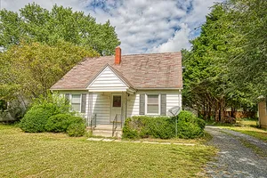 Charming single-story home with a cozy front porch, nestled amid lush greenery and mature trees, featuring a classic gable roof.