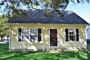 Charming yellow cottage with dark shutters and a welcoming front door, surrounded by lush green lawn and shaded by trees.