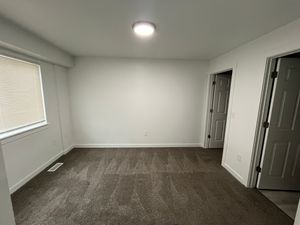 Bright and spacious room with clean carpet, fresh white walls, and ample natural light from a large window, featuring two doors for versatile access.
