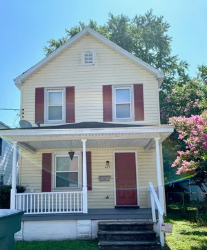 Charming two-story house with a welcoming front porch, vibrant red shutters, and lush greenery, perfect for a cozy family home.
